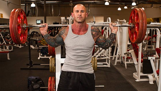 an inked up bodybuilder holding a barbell across his shoulders