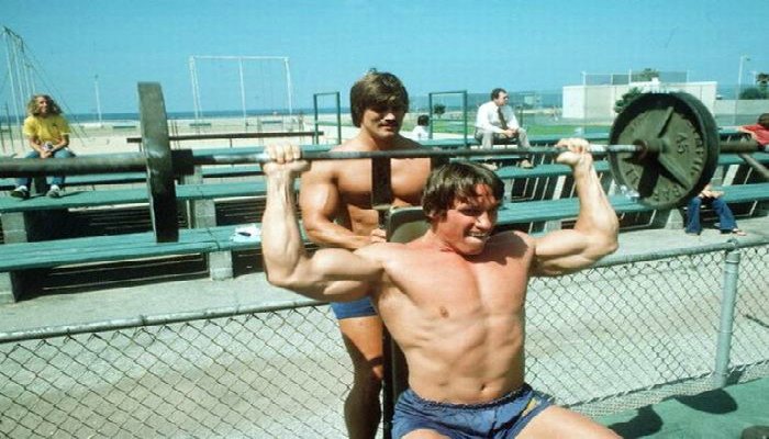 Arnold and Frank Columbo lifting weights for shoulders, working out on the beach