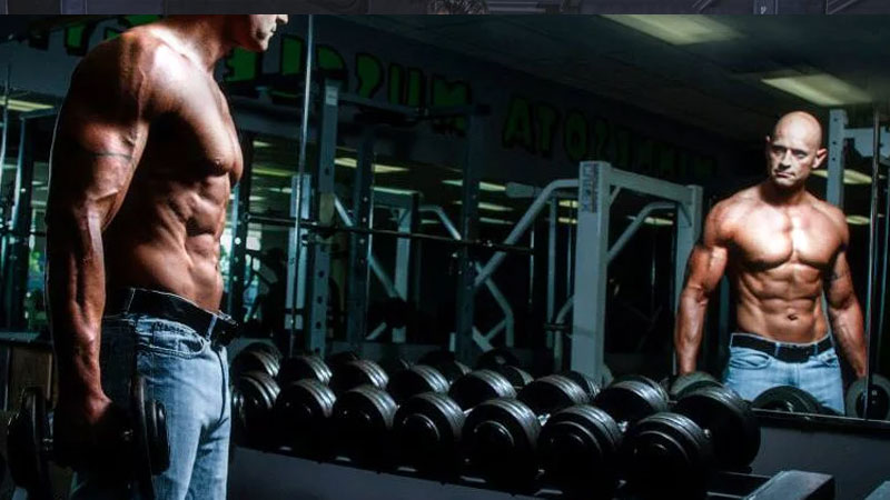 Bodybuilder poses in front of a mirror in the gym with dumbbells