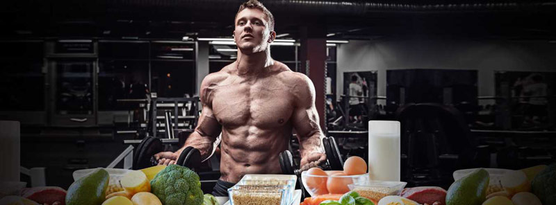 bodybuilder holding dumbbells standing behind row of nutritious whole foods to build muscle without taking any supplements